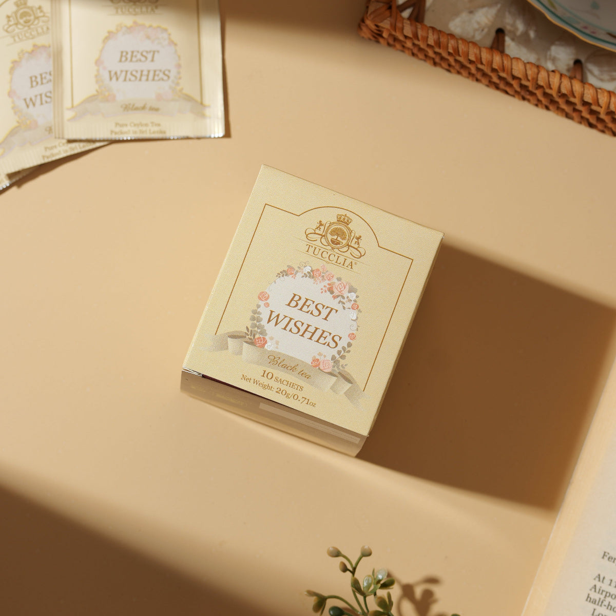 Box of 'Best Wishes' tea bags on a beige surface with a cup of tea and flowers in the background.